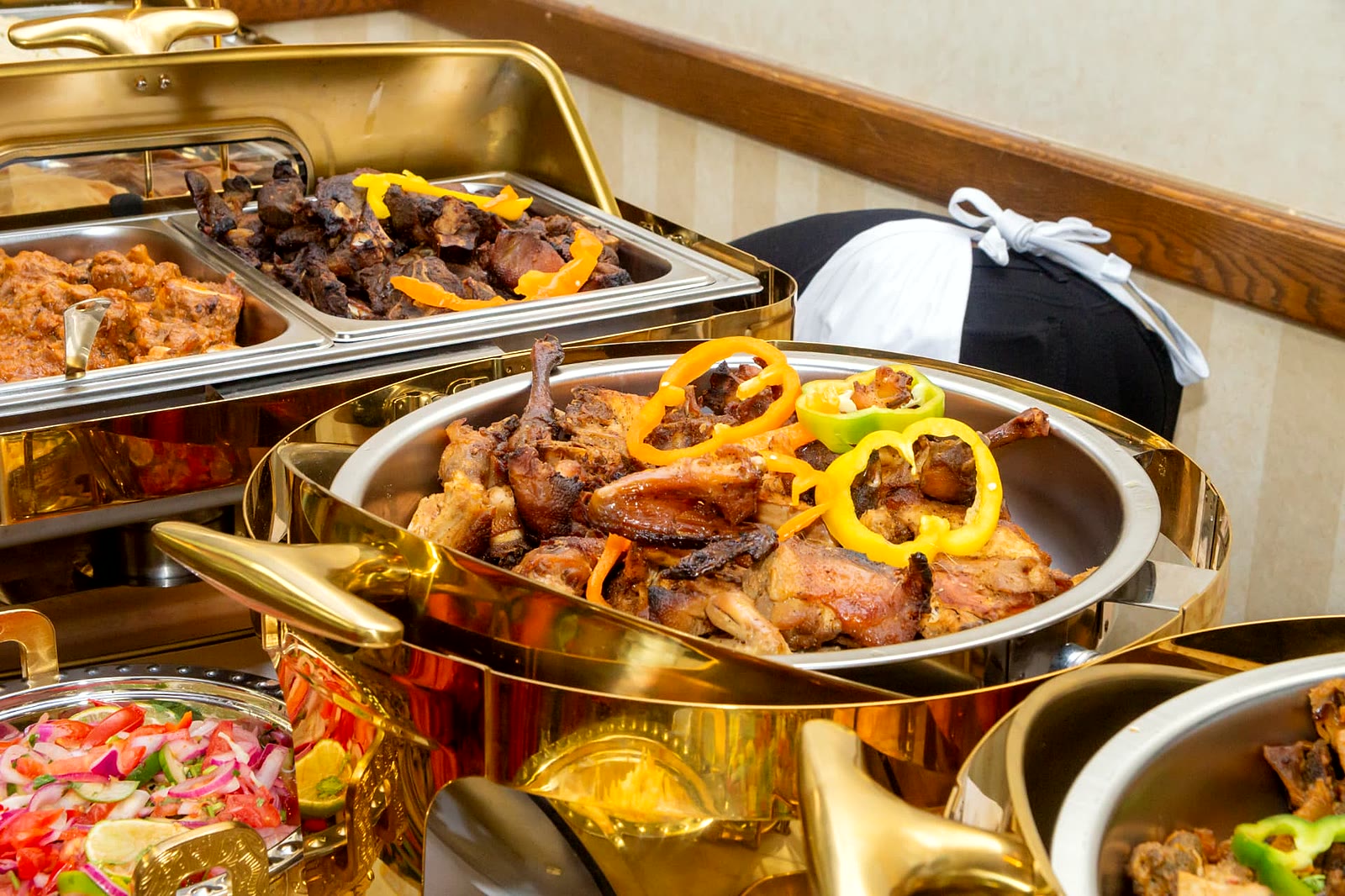 Roasted goat meat and grilled chicken garnished with peppers in gold chafing dishes at a Toronto event